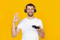 年轻的男人。胡子白色t恤玩家玩视频游戏耳机使正常的标志手指优秀的象征站孤立的黄色的背景