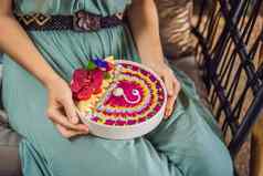 年轻的女人地中海早餐坐着沙发吃健康的热带早餐奶昔碗热带水果装饰模式色彩斑斓的酸奶姜黄螺旋藻装饰水果花传讲种子椰子格兰诺拉麦片菠萝薄荷草莓