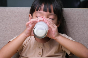 肖像可爱的亚洲女孩持有玻璃牛奶坐着沙发<strong>首页</strong>小女孩<strong>首页</strong>微笑脸感觉快乐享受喝牛奶相机