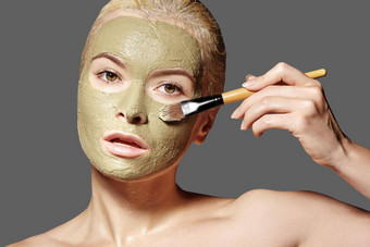 美丽的女人应用绿色面部面具美治疗水疗中心女孩应用粘土面部面具灰色背景
