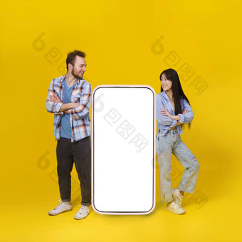 亚洲女孩高加索人的家伙站靠<strong>巨</strong>大的智能手机白色空白屏<strong>幕</strong>移动应用程序广告微笑孤立的黄色的背景产品放置