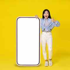 兴奋亚洲女孩手势拇指靠巨大的巨大的智能手机白色空白屏幕站靠穿休闲孤立的黄色的背景免费的空间模拟