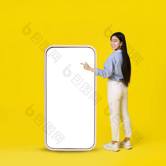 亚洲女孩指出巨大的巨大的智能<strong>手机</strong>白色空白屏幕站回来快乐指出手指穿着休闲孤立的黄色的背景免费的空间<strong>模拟</strong>