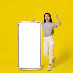 快乐亚洲女孩靠巨大的巨大的智能手机白色空白屏幕快乐指出手指穿着休闲孤立的黄色的背景免费的空间模拟