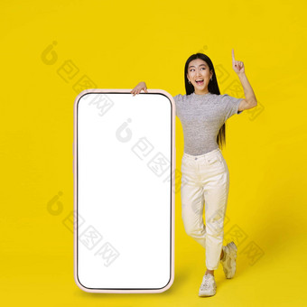 快乐亚洲女孩靠巨大的巨大的智能手机白色空白屏幕快乐指出手指穿着休闲孤立的黄色的背景免费的空间模拟