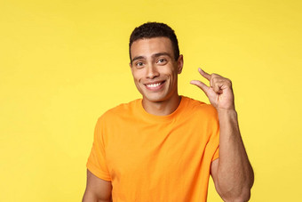 快乐的英俊的微笑男人。橙色t恤显示小手势咧着嘴笑很高兴小请求手势对象站满意黄色的背景