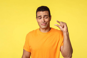 持怀疑态度失望年轻的英俊的男人。橙色t恤显示小讨论小对象得意的笑鄙视手指不站黄色的背景