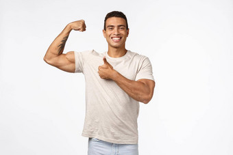 英俊的强大的运动运动员休闲t恤提高手臂紧张的肌肉自豪地吹牛好形状大肱二头肌微笑很高兴显示竖起大拇指批准白色背景