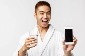 美水疗中心休闲概念快乐年轻的亚洲男人。浴袍促进智能手机应用程序检查医疗保健显示移动电话屏幕持有玻璃水