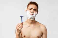 美人卫生概念不情愿的不高兴亚洲男人。不愿刮胡子失望相机扮鬼脸显示剃须刀应用奶油下颌的轮廓白色背景