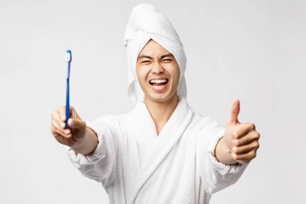 美水疗中心休闲概念肖像热情的英俊的亚洲男人。浴袍浴毛巾显示牙刷满意脸使翘拇指阿波瓦尔推荐医疗保健产品