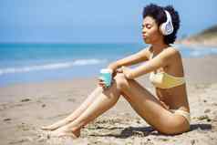 非洲美国女孩坐着海滩耳机喝着喝外卖杯