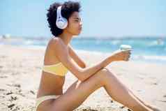 黑色的女人坐着海滩耳机喝着喝纸外卖杯