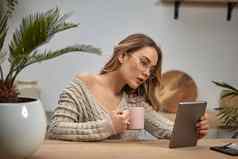女人博主眼镜米色毛衣持有粉红色的杯平板电脑坐着厨房木表格棕榈智能手机塑料卡特写镜头