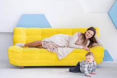 年轻的快乐高加索人女人谎言黄色的沙发手表婴儿爬地板上等待婴儿的步骤