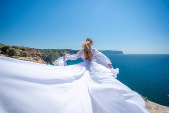 金发女郎长头发阳光明媚的海滨白色流动衣服后视图丝绸织物挥舞着风背景蓝色的天空山海滨