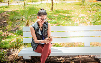 女孩坐着板凳上检查细胞电话快乐女人坐着公园发短信细胞电话女人坐着板凳上发送文本消息