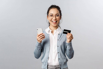 快乐的年轻的亚洲女人<strong>推荐</strong>银行订单在线使互联网购买交付应用程序持有信贷卡智能手机微笑满意灰色背景