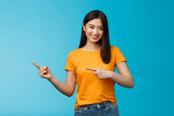 友好的即将离任的好看的亚洲女人显示促销指出左指示手指广告给建议微笑自信愉快地选择衣服站蓝色的背景