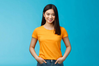 自信自信的微笑亚洲女人站蓝色的背景相机持有手牛仔裤口袋表达自信的氛围享受积极的结果护肤品程序穿黄色的t恤