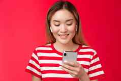 无忧无虑的现代亚洲金发碧眼的女孩挑选首歌穿无线耳机听音乐在线首歌平台应用程序持有智能手机很高兴微笑电话屏幕站红色的背景