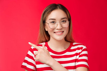 好机会角落里可爱的亚洲女孩穿眼镜条纹夏天t恤指出左微笑露出牙齿的友好的表达式女人介绍促销指示好选择红色的背景