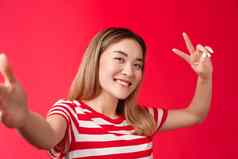 友好的即将离任的可爱的亚洲女孩金发碧眼的短发型扩展手臂持有智能手机相机倾斜头快乐显示和平胜利标志采取自拍拍摄站红色的背景