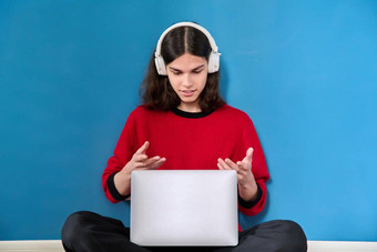 十几岁的男孩学生耳机移动PC会议视频调用坐着地板上蓝色的背景