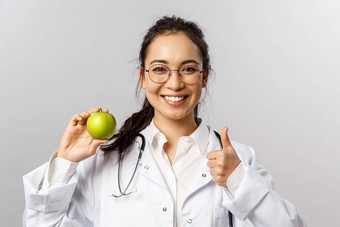 肖像年轻的快乐的亚洲女医生治疗师diatologist会说话的邻接健康的饮食显示绿色苹果翘拇指推荐吃水果微笑鼓励健康
