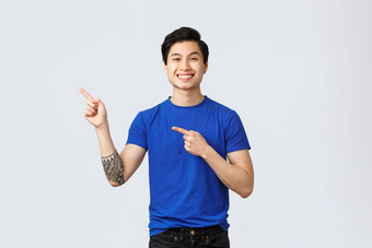 英俊的年轻的亚洲男人。蓝色的t恤纹身手臂指出手指上左角落里微笑介绍广告产品站灰色背景快乐的