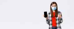 情绪科维德社会距离技术概念兴奋年轻的快乐女人推荐好应用程序显示移动电话屏幕翘拇指穿医疗面具