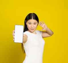 自信美丽的亚洲女人微笑持有智能手机显示空白屏幕移动应用程序广告微笑相机孤立的黄色的背景产品放置