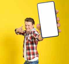 快乐赢得英俊的男人。指出智能手机白色空屏幕穿红色的格子衬衫牛仔裤手机显示模拟孤立的黄色的背景移动应用程序广告