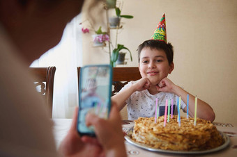 快乐的高加索人十几岁的男孩坐着表格生日蛋糕蜡烛微笑摆姿势模糊妈妈采取照片智能手机前景生日聚会，派对概念