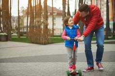 英俊的欧洲爱的有爱心的爸爸走女儿骑踢踏板车走城市公园快乐家庭的关系无忧无虑的童年概念