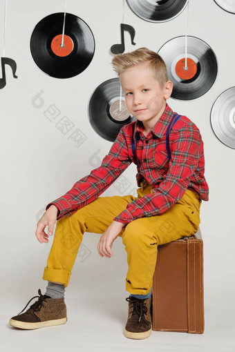 复古的迪斯科概念有趣的男孩穿检查衬衫时尚的发型坐着手提箱背景音乐板