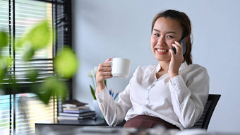 微笑亚洲女人持有咖啡杯享受电话谈话工作场所