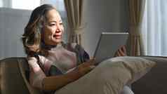 退休女人坐着舒适的沙发检查电子邮件新闻在线平板电脑上了年纪的技术概念