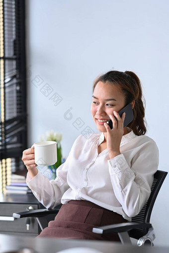 微笑亚洲女人持有咖啡杯会说话的手机办公室桌子上