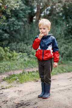 肖像男孩显示鬼脸孩子色彩斑斓的夹克穿防水靴子站边缘森林