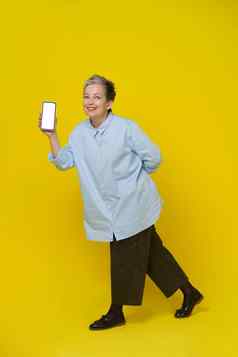 显示电话手成熟的灰色头发的女人微笑相机漂亮的女人蓝色的衬衫棕色（的）裤子孤立的黄色的背景移动应用程序广告模型产品放置