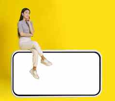 深思熟虑的亚洲女孩坐巨大的智能手机白色屏幕穿休闲白色牛仔裤孤立的黄色的背景模拟产品放置移动应用程序广告复制空间