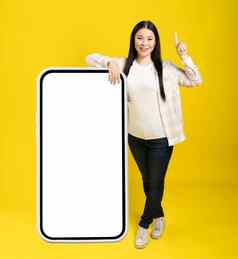亚洲女人指出手指注意靠回来巨大的巨大的智能手机空白白色屏幕穿休闲孤立的黄色的背景免费的空间模拟