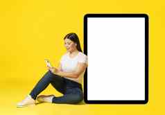 亚洲女人靠巨大的巨大的平板电脑坐着地板上电话手休闲白色屏幕电话移动应用程序广告孤立的黄色的产品放置