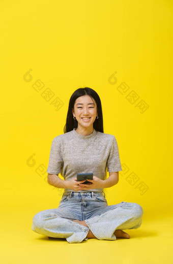 温柔的亚洲年轻的女孩坐着地板上电话手发短信购物在线玩游戏孤立的黄色的背景移动应用程序广告复制空间