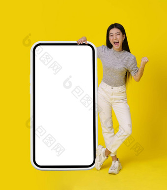 完整的身体亚洲女孩站巨大的巨大的智能手机白色屏幕快乐使是的赢家手手势穿着休闲孤立的黄色的背景免费的空间模拟