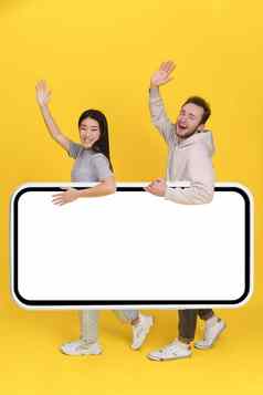 问候英俊的的家伙亚洲女孩持有巨大的智能手机数字平板电脑白色屏幕移动应用程序广告兴奋微笑相机孤立的黄色的背景产品放置