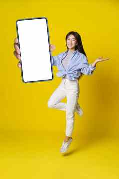亚洲女孩跳持有智能手机黑色的屏幕手休闲装移动应用程序广告孤立的黄色的背景完整的长度肖像快乐的亚洲女孩产品放置