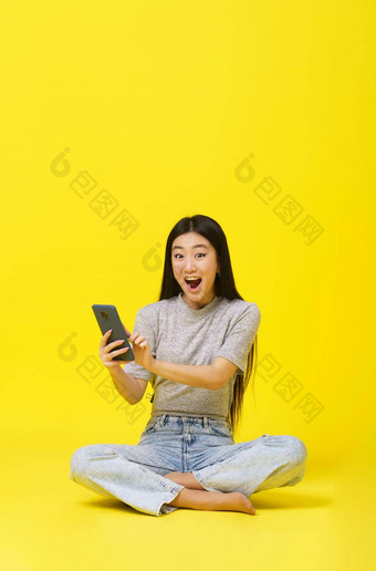 兴奋亚洲年轻的女孩坐着地板上持有智能手机发短信购物在线赢得游戏孤立的黄色的背景产品放置移动应用程序广告复制空间