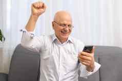 快乐兴奋祖父持有智能手机笑大喊大叫快乐好新闻阅读文本消息屏幕使赢家手手势惊讶老男人。赢得奖
